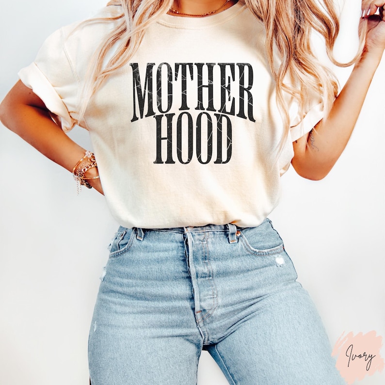 May include: A white t-shirt with the text "MOTHERHOOD" printed in black on the front. The shirt is being worn by a person with long blonde hair and blue jeans.