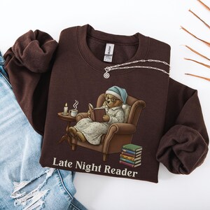 Gildan 18000 sweatshirt featuring a cozy teddy bear wearing a nightgown and hat, sitting in a chair reading a book with a candle, coffee cup, and stack of books beside him. The design includes the text “Late Night Reader” beneath the image. Styled in a warm, vintage-inspired look perfect for book lovers, cozy fall nights, and readers who enjoy a comforting, bookish aesthetic.