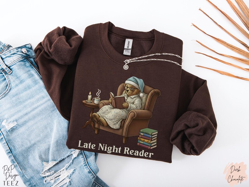 Gildan 18000 sweatshirt featuring a cozy teddy bear wearing a nightgown and hat, sitting in a chair reading a book with a candle, coffee cup, and stack of books beside him. The design includes the text “Late Night Reader” beneath the image. Styled in a warm, vintage-inspired look perfect for book lovers, cozy fall nights, and readers who enjoy a comforting, bookish aesthetic.
