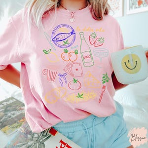 K&ouml;nnte beinhalten: Hellrosa T-Shirt mit farbenfrohen Illustrationen italienischer Speisen und Getr&auml;nke, darunter ein Fisch, eine Weinflasche und Erdbeeren. Die Worte "la dolce vita" sind in Schrift geschrieben. Eine wei&szlig;e Tasse mit einem gelben Smiley wird gehalten.