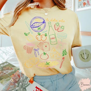 K&ouml;nnte beinhalten: Hellgelbes T-Shirt mit bunten Illustrationen italienischer Speisen und Getr&auml;nke, darunter ein Fisch, eine Weinflasche, Erdbeeren und Pasta. Die Worte "la dolce vita" und "ciao" sind ebenfalls abgebildet. Eine Tasse mit einem Smiley ist sichtbar.