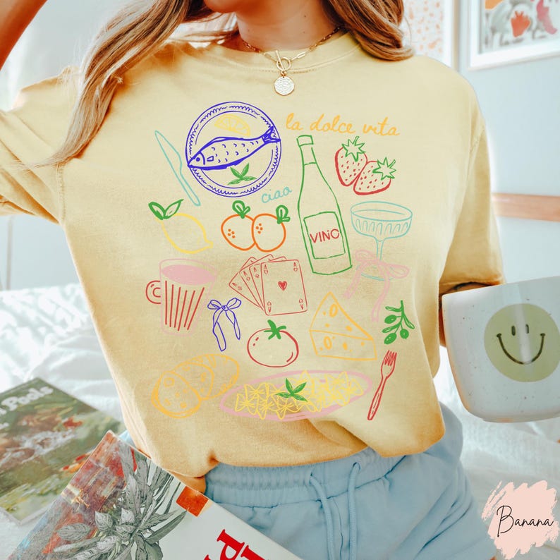 K&ouml;nnte beinhalten: Hellgelbes T-Shirt mit bunten Illustrationen italienischer Speisen und Getr&auml;nke, darunter ein Fisch, eine Weinflasche, Erdbeeren und Pasta. Die Worte "la dolce vita" und "ciao" sind ebenfalls abgebildet. Eine Tasse mit einem Smiley ist sichtbar.