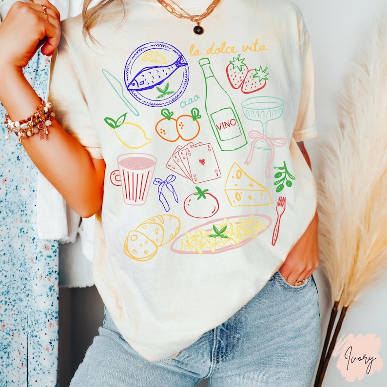 K&ouml;nnte beinhalten: Cremefarbenes T-Shirt mit bunten Illustrationen italienischer Speisen und Getr&auml;nke. Das Design umfasst einen Fisch, eine Weinflasche, Erdbeeren und die Worte "la dolce vita" und "ciao". Das Shirt ist ein l&auml;ssiger Stil.