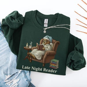 Gildan 18000 sweatshirt featuring a cozy teddy bear wearing a nightgown and hat, sitting in a chair reading a book with a candle, coffee cup, and stack of books beside him. The design includes the text “Late Night Reader” beneath the image. Styled in a warm, vintage-inspired look perfect for book lovers, cozy fall nights, and readers who enjoy a comforting, bookish aesthetic.