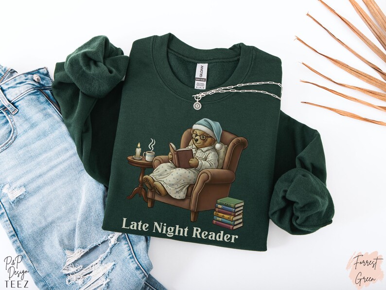 Gildan 18000 sweatshirt featuring a cozy teddy bear wearing a nightgown and hat, sitting in a chair reading a book with a candle, coffee cup, and stack of books beside him. The design includes the text “Late Night Reader” beneath the image. Styled in a warm, vintage-inspired look perfect for book lovers, cozy fall nights, and readers who enjoy a comforting, bookish aesthetic.