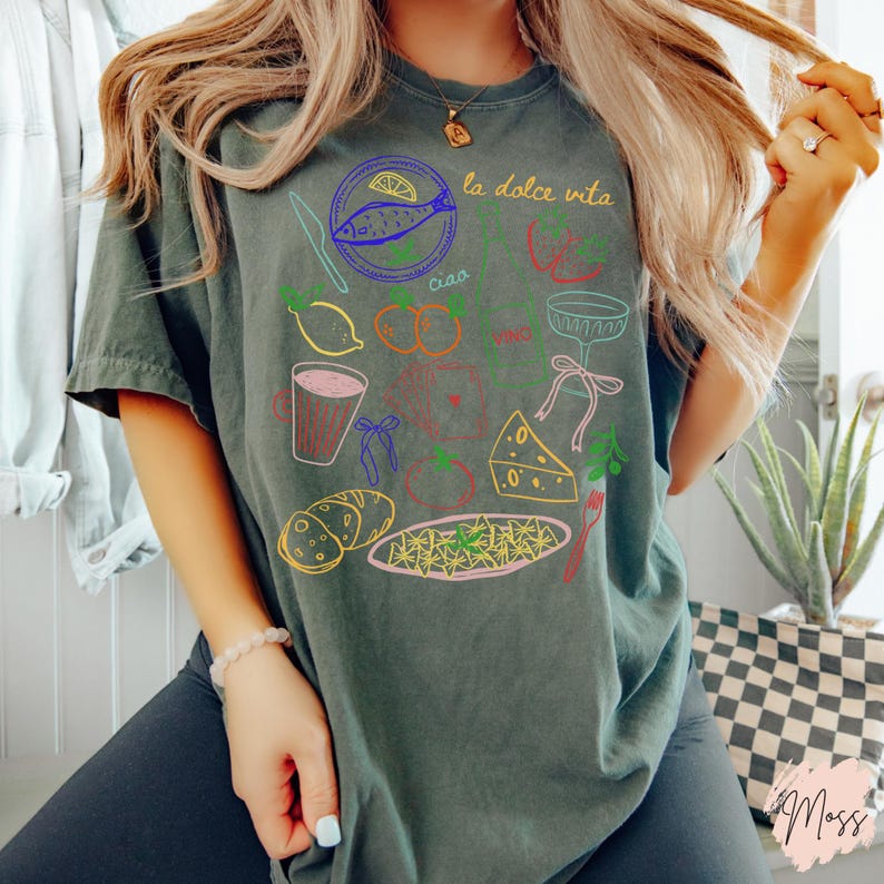 K&ouml;nnte beinhalten: Dunkelgr&uuml;nes T-Shirt mit einer farbenfrohen Grafik zum Thema italienisches Essen. Das Design umfasst Illustrationen von Fisch, Zitronen, Tomaten, Pasta und die Worte "la dolce vita" und "ciao". Das Shirt ist ein l&auml;ssiger, bequemer Stil.