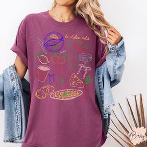 K&ouml;nnte beinhalten: Ein weinrotes T-Shirt mit einem farbenfrohen Grafikdesign mit italienischem Essen und den Worten "la dolce vita" und "ciao". Das Design umfasst einen Fisch, Zitronen, Tomaten, Pasta und ein Glas.