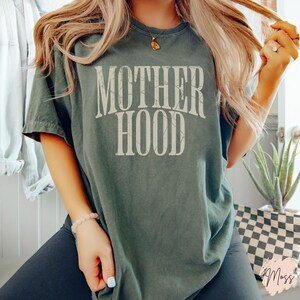 May include: A dark green t-shirt with the text "MOTHERHOOD" printed in white on the front. The text has a distressed, vintage look.