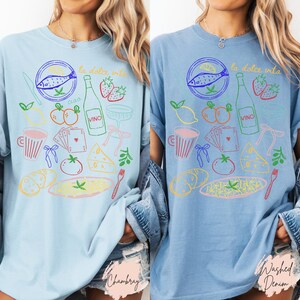 K&ouml;nnte beinhalten: Hellblaue T-Shirts mit einem farbenfrohen italienischen Food-Themen-Grafik. Das Design umfasst einen Fisch, eine Weinflasche, Erdbeeren und andere Lebensmittel, mit dem Text "la dolce vita".