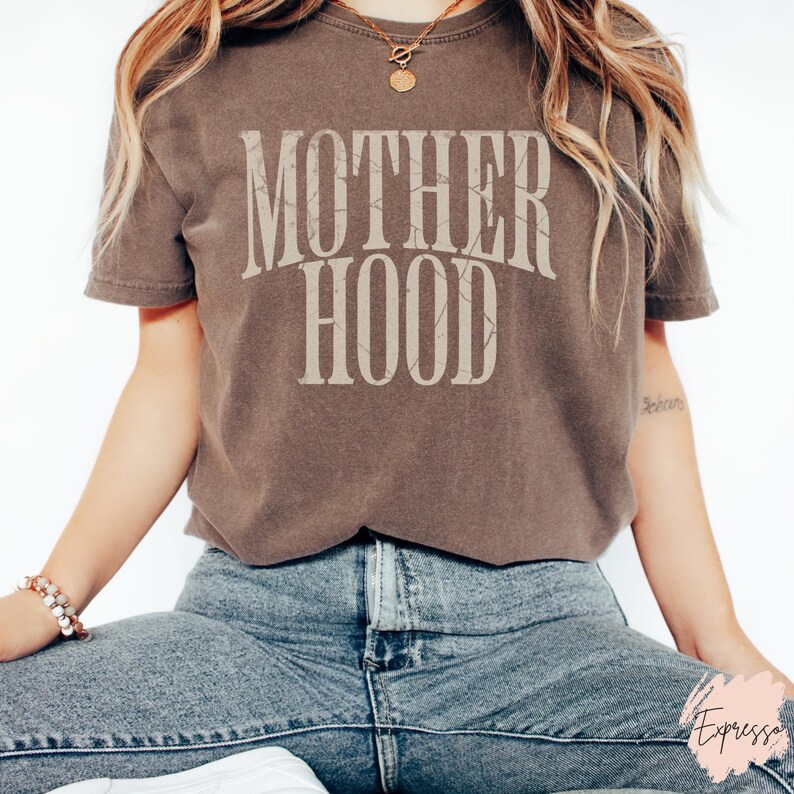 May include: A brown t-shirt with the text "MOTHERHOOD" printed in white on the front. The shirt is being worn by a person with long brown hair and is styled with blue jeans.