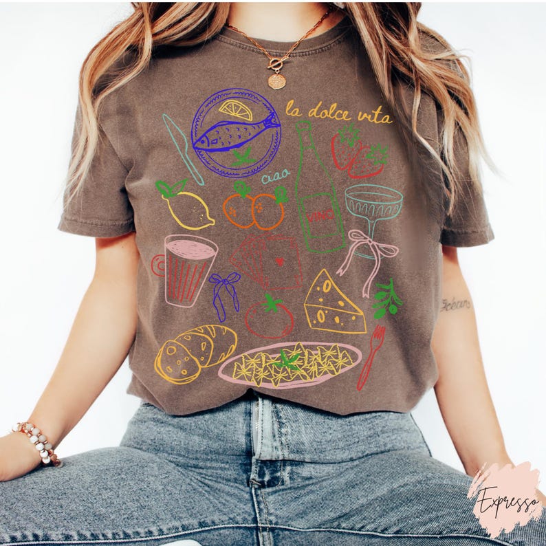 K&ouml;nnte beinhalten: Braunes T-Shirt mit farbenfroher Strichzeichnung italienischer Speisen und Ausdr&uuml;cke. Das Design umfasst einen Fisch, Zitronen, Tomaten, Pasta, K&auml;se und die Worte "la dolce vita" und "ciao".