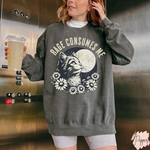 Embrace your feral energy with this distressed effect vintage-inspired Rage Consumes Me funny cat sweatshirt, printed on the ultra-comfy Comfort Colors 1566 sweatshirt. Designed for a relaxed, oversized fit, this unisex crewneck is perfect for cat lovers, introverts, and anyone who thrives on a little chaos. Featuring a moody cat, full moon, and floral details, this design brings together cottagecore and grunge aesthetics with a touch of humor.