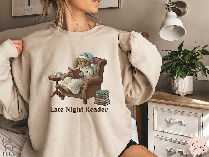 Gildan 18000 sweatshirt featuring a cozy teddy bear wearing a nightgown and hat, sitting in a chair reading a book with a candle, coffee cup, and stack of books beside him. The design includes the text “Late Night Reader” beneath the image. Styled in a warm, vintage-inspired look perfect for book lovers, cozy fall nights, and readers who enjoy a comforting, bookish aesthetic.