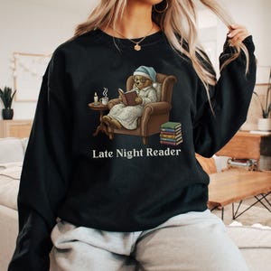 Gildan 18000 sweatshirt featuring a cozy teddy bear wearing a nightgown and hat, sitting in a chair reading a book with a candle, coffee cup, and stack of books beside him. The design includes the text “Late Night Reader” beneath the image. Styled in a warm, vintage-inspired look perfect for book lovers, cozy fall nights, and readers who enjoy a comforting, bookish aesthetic.