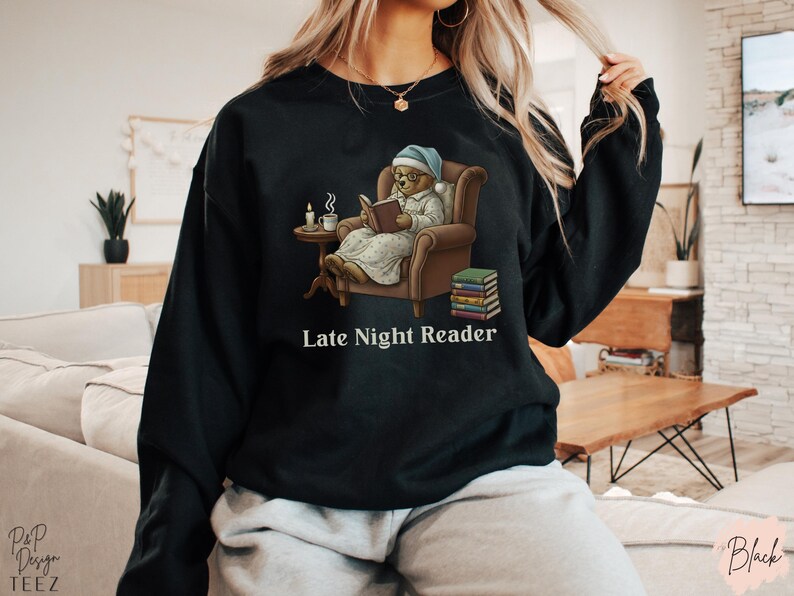 Gildan 18000 sweatshirt featuring a cozy teddy bear wearing a nightgown and hat, sitting in a chair reading a book with a candle, coffee cup, and stack of books beside him. The design includes the text “Late Night Reader” beneath the image. Styled in a warm, vintage-inspired look perfect for book lovers, cozy fall nights, and readers who enjoy a comforting, bookish aesthetic.