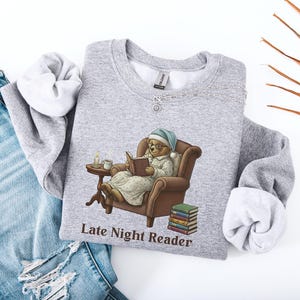 Gildan 18000 sweatshirt featuring a cozy teddy bear wearing a nightgown and hat, sitting in a chair reading a book with a candle, coffee cup, and stack of books beside him. The design includes the text “Late Night Reader” beneath the image. Styled in a warm, vintage-inspired look perfect for book lovers, cozy fall nights, and readers who enjoy a comforting, bookish aesthetic.