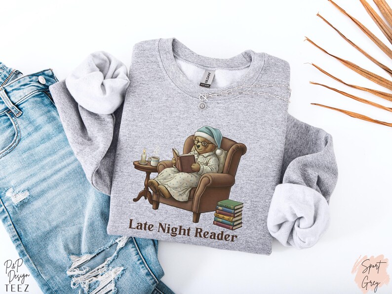 Gildan 18000 sweatshirt featuring a cozy teddy bear wearing a nightgown and hat, sitting in a chair reading a book with a candle, coffee cup, and stack of books beside him. The design includes the text “Late Night Reader” beneath the image. Styled in a warm, vintage-inspired look perfect for book lovers, cozy fall nights, and readers who enjoy a comforting, bookish aesthetic.
