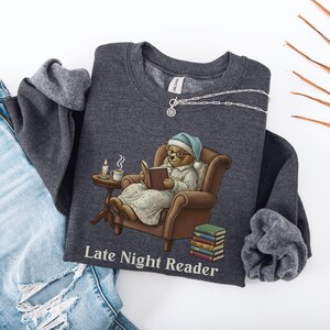 Gildan 18000 sweatshirt featuring a cozy teddy bear wearing a nightgown and hat, sitting in a chair reading a book with a candle, coffee cup, and stack of books beside him. The design includes the text “Late Night Reader” beneath the image. Styled in a warm, vintage-inspired look perfect for book lovers, cozy fall nights, and readers who enjoy a comforting, bookish aesthetic.