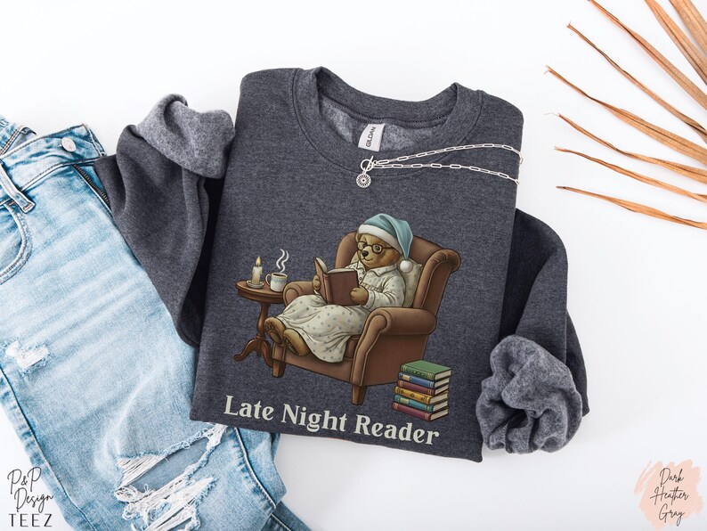 Gildan 18000 sweatshirt featuring a cozy teddy bear wearing a nightgown and hat, sitting in a chair reading a book with a candle, coffee cup, and stack of books beside him. The design includes the text “Late Night Reader” beneath the image. Styled in a warm, vintage-inspired look perfect for book lovers, cozy fall nights, and readers who enjoy a comforting, bookish aesthetic.