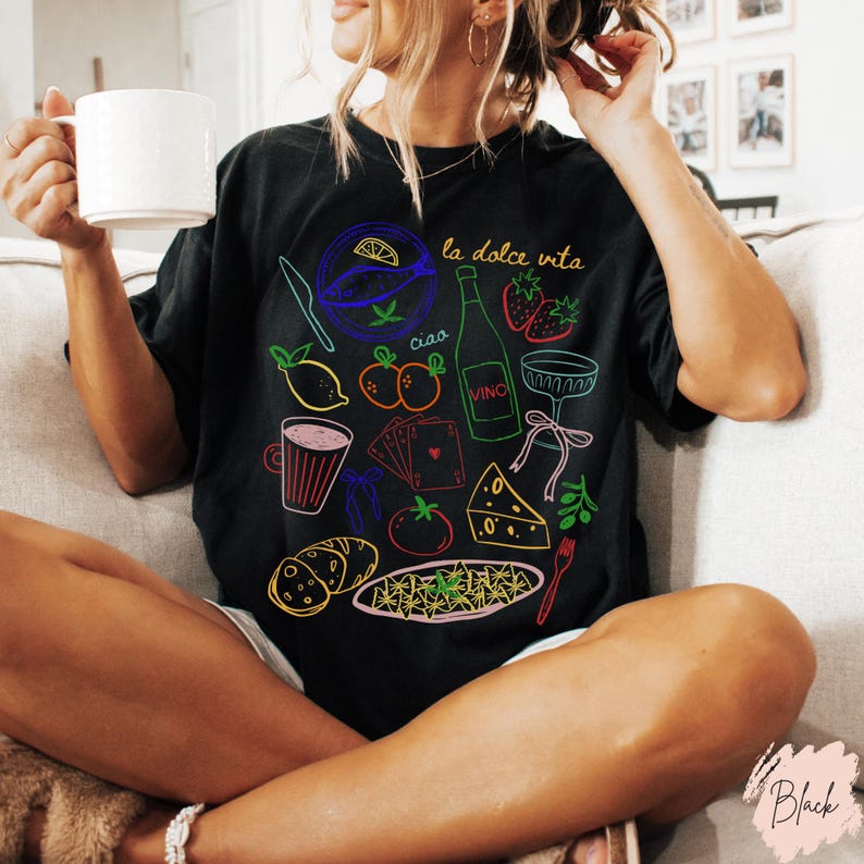 K&ouml;nnte beinhalten: Schwarzes T-Shirt mit farbenfroher Strichzeichnung italienischer Speisen und Getr&auml;nke, darunter eine Weinflasche, Erdbeeren und K&auml;se. Die Worte "la dolce vita" und "ciao" sind ebenfalls auf dem Shirt zu sehen.