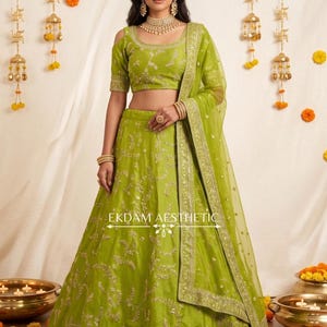 Puede incluir: Un conjunto de lehenga choli verde lima con intrincados bordados dorados, escote redondo, mangas con hombros descubiertos y una dupatta transparente a juego. La falda tiene un borde ancho bordado. El texto EKDAM AESTHETIC es visible en la falda.