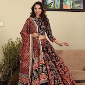 Puede incluir: Una mujer con un atuendo tradicional indio. El conjunto incluye un top y una falda con estampado negro y rojo, combinados con un chal a juego. El atuendo presenta diseños intrincados y se encuentra sobre un fondo neutro.