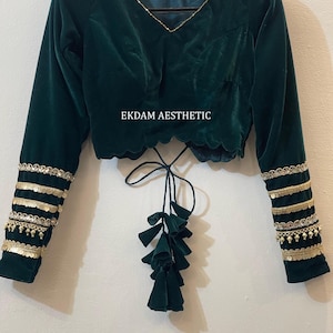 May include: A dark green velvet blouse with long sleeves and a scalloped hem. The sleeves are adorned with gold trim and embellishments. The blouse has a V-neckline with gold detailing and a tie closure with tassels. The text "EKDAM AESTHETIC" is printed on the blouse.
