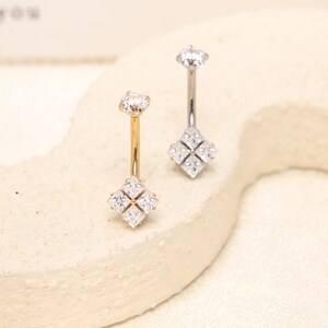 Pode incluir: Dois piercings de umbigo com design de barra curva. Um é de tom dourado e o outro de tom prateado. Cada peça é rematada por uma pedra redonda transparente e termina com um elemento quadrado composto por quatro pedras transparentes.