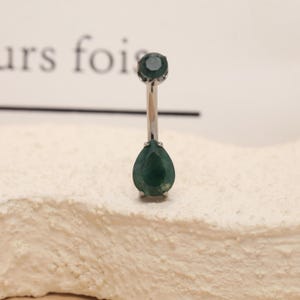 May include: A silver belly button ring with two dark green, teardrop-shaped gemstones. The top gem is round, and the bottom gem is faceted. The ring is displayed on a textured, off-white surface.
