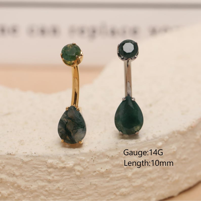 May include: Two belly button rings with teardrop-shaped green gemstones. One ring has a gold-colored curved bar, and the other has a silver-colored bar. The text "Gauge: 14G Length: 10mm" is visible.