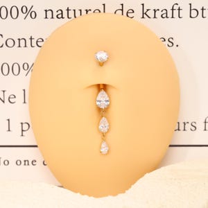 Pode incluir: Um piercing de umbigo dourado com uma pedra preciosa redonda e transparente no topo e uma corrente vertical pendente de tr&ecirc;s pedras transparentes em forma de pera de tamanho graduado, exibido num modelo oval bege de um umbigo humano.