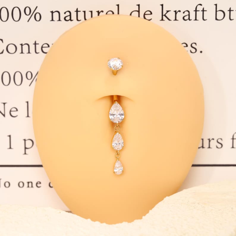Pode incluir: Um piercing de umbigo dourado com uma pedra preciosa redonda e transparente no topo e uma corrente vertical pendente de tr&ecirc;s pedras transparentes em forma de pera de tamanho graduado, exibido num modelo oval bege de um umbigo humano.