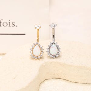 Pode incluir: Dois anéis de umbigo com uma pedra central de opala branca em forma de lágrima rodeada por um halo de pequenos cristais redondos transparentes. Um anel tem uma barra curva dourada e o outro uma prateada, ambos com um cristal no topo.