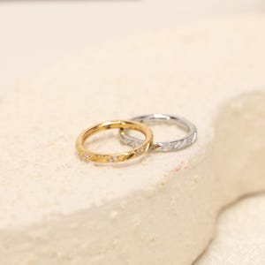 May include: Two circular hoop rings, one in a gold tone and one in a silver tone, featuring a textured surface with small embedded clear stones. The rings are thin, metallic, and designed with a hinged closure mechanism for secure fastening.