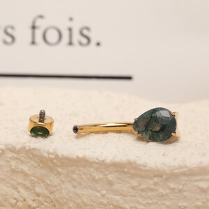 May include: A gold-colored body piercing jewelry with a teardrop-shaped green gemstone. The jewelry is in two pieces, with a threaded post and a decorative end. The gemstone has a unique pattern.