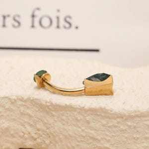 May include: A gold-colored belly button ring with two green gemstones. One gemstone is round, and the other is square. The ring has a curved bar and is set against a textured, off-white surface.