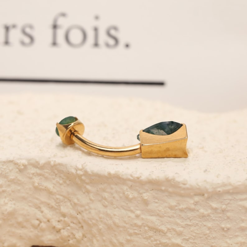 May include: A gold-colored belly button ring with two green gemstones. One gemstone is round, and the other is square. The ring has a curved bar and is set against a textured, off-white surface.