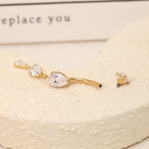 Pode incluir: Um piercing de umbigo curvo em tom dourado com uma corrente pendente de tr&ecirc;s pedras de zirc&oacute;nia c&uacute;bica transparentes em forma de pera. A pe&ccedil;a inclui uma bola superior aparafus&aacute;vel separada com uma pedra transparente e acabamento met&aacute;lico.