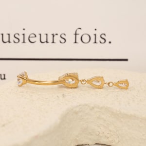 Pode incluir: Um piercing de umbigo curvo em tom dourado com uma pedra preciosa redonda transparente numa extremidade e um pendente com duas pedras transparentes em forma de pera ligadas por pequenos elos dourados.