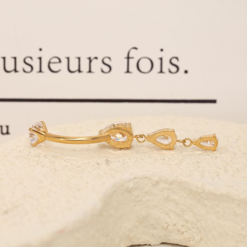 Pode incluir: Um piercing de umbigo curvo em tom dourado com uma pedra preciosa redonda transparente numa extremidade e um pendente com duas pedras transparentes em forma de pera ligadas por pequenos elos dourados.