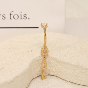 Pode incluir: Um piercing de umbigo de barra curva em tom dourado com uma esfera superior e um design pendente vertical. O pendente consiste em tr&ecirc;s pedras transparentes em forma de pera cravadas em metal dourado, dispostas numa linha descendente.