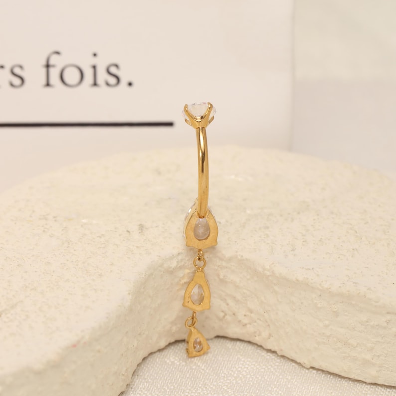 Pode incluir: Um piercing de umbigo de barra curva em tom dourado com uma esfera superior e um design pendente vertical. O pendente consiste em tr&ecirc;s pedras transparentes em forma de pera cravadas em metal dourado, dispostas numa linha descendente.