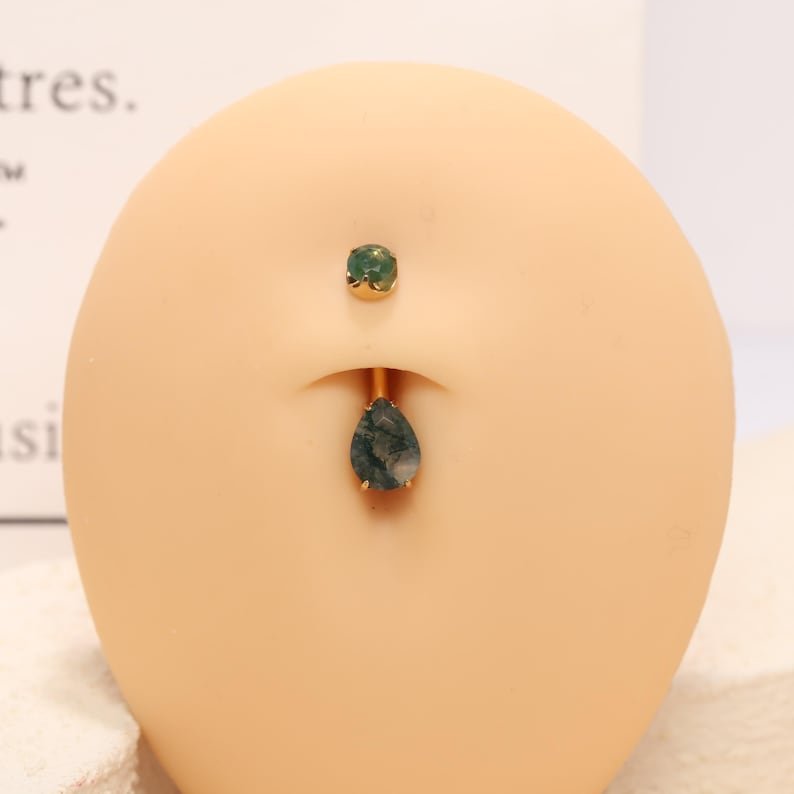 May include: Gold-toned belly button ring with a teardrop-shaped green stone pendant. The top stud is a small, round green stone. The jewelry is displayed on a light-colored surface.