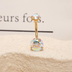Pode incluir: Um piercing no umbigo dourado com duas pedras preciosas redondas e iridescentes. A gema maior está na parte inferior, e uma menor, em forma de gota, está na parte superior. A joia está sobre um fundo neutro.