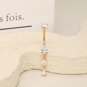May include: A gold-toned curved barbell belly ring featuring a round clear gemstone at the top and three dangling pear-shaped clear gemstones arranged vertically. The jewellery has a polished metallic finish and clear faceted stones.