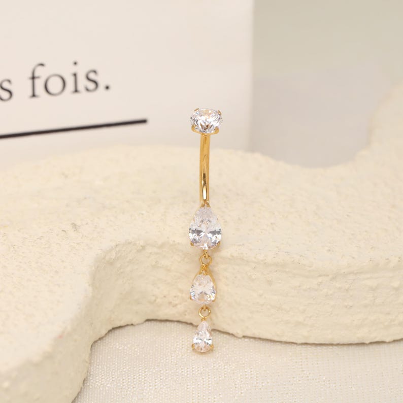 Pode incluir: Um piercing de umbigo curvo em tom dourado com uma pedra preciosa redonda transparente no topo e tr&ecirc;s pedras transparentes em forma de pera penduradas verticalmente. A joia tem um acabamento met&aacute;lico polido e pedras facetadas.