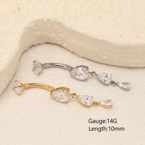 Pode incluir: Dois piercings curvos para umbigo, um prateado e outro dourado, com calibre 14G e comprimento de 10mm. Ambas as pe&ccedil;as possuem um cristal transparente em forma de pera em cada extremidade, com um pendente contendo dois cristais adicionais.