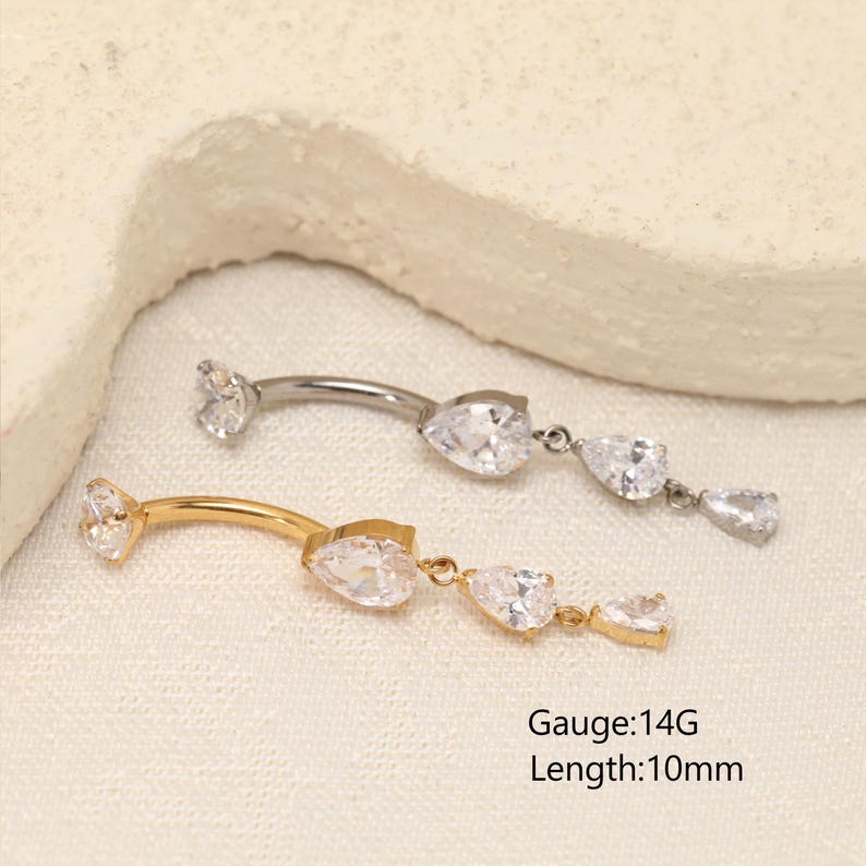 Pode incluir: Dois piercings curvos para umbigo, um prateado e outro dourado, com calibre 14G e comprimento de 10mm. Ambas as pe&ccedil;as possuem um cristal transparente em forma de pera em cada extremidade, com um pendente contendo dois cristais adicionais.
