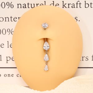 Pode incluir: Um piercing de umbigo prateado com uma pedra transparente redonda no topo e uma corrente vertical pendente composta por tr&ecirc;s pedras transparentes graduadas em forma de pera. A joia &eacute; exibida num modelo anat&oacute;mico de cor bege.