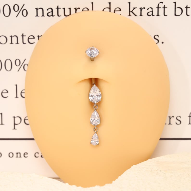 Pode incluir: Um piercing de umbigo prateado com uma pedra transparente redonda no topo e uma corrente vertical pendente composta por tr&ecirc;s pedras transparentes graduadas em forma de pera. A joia &eacute; exibida num modelo anat&oacute;mico de cor bege.